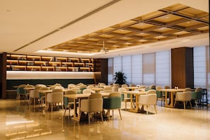A spacious dining area with multiple tables and chairs, a modern ceiling design, and a large bookshelf.
