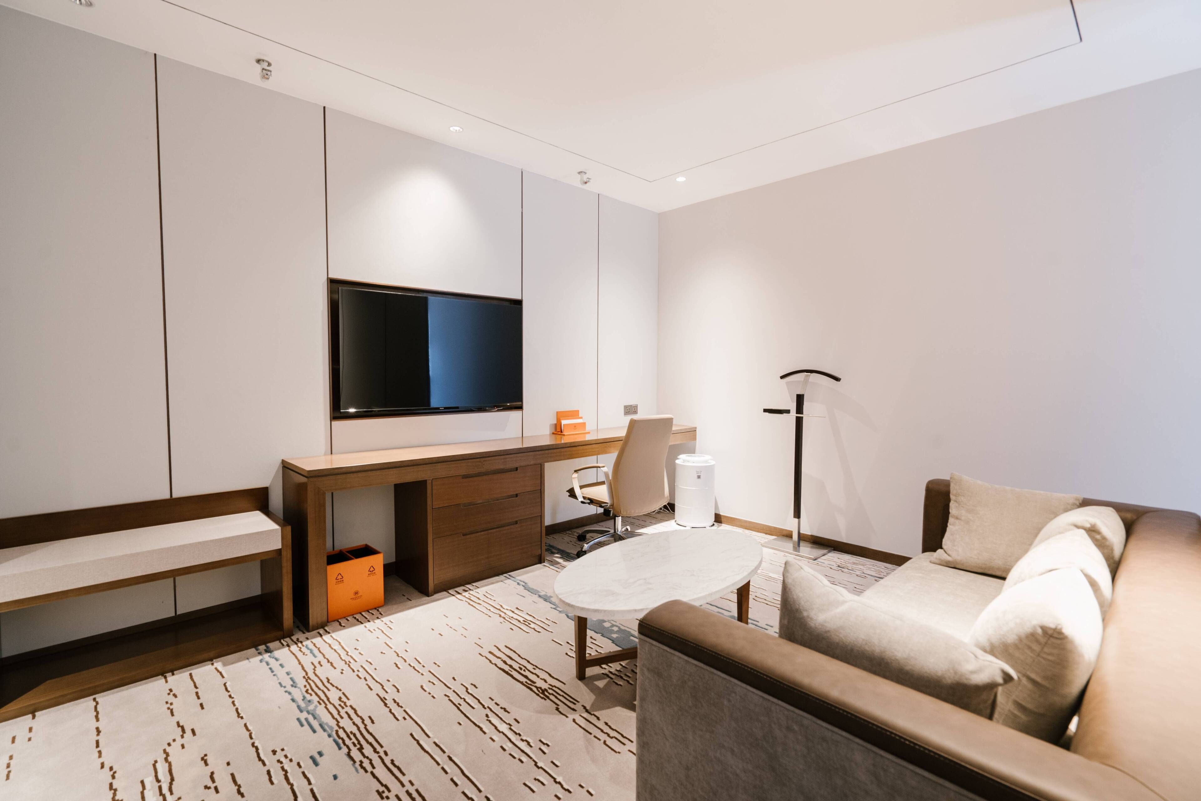 A modern hotel room with a flat-screen TV mounted on the wall, a wooden desk with drawers, a sofa, a coffee table, and a floor lamp.