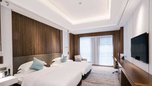 Ein Hotelzimmer mit zwei Betten, einem Schreibtisch, einem Sessel, einem Fernseher und einem großen Fenster.