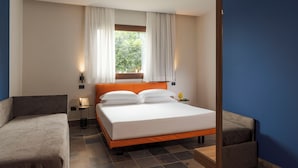 Habitación de hotel con cama, dos sillones, una mesita de noche con lámpara y una ventana con cortinas.