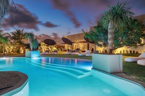 Ein Pool mit Liegestühlen und Sonnenschirmen, umgeben von Palmen und einem Gebäude mit Steinmauern.