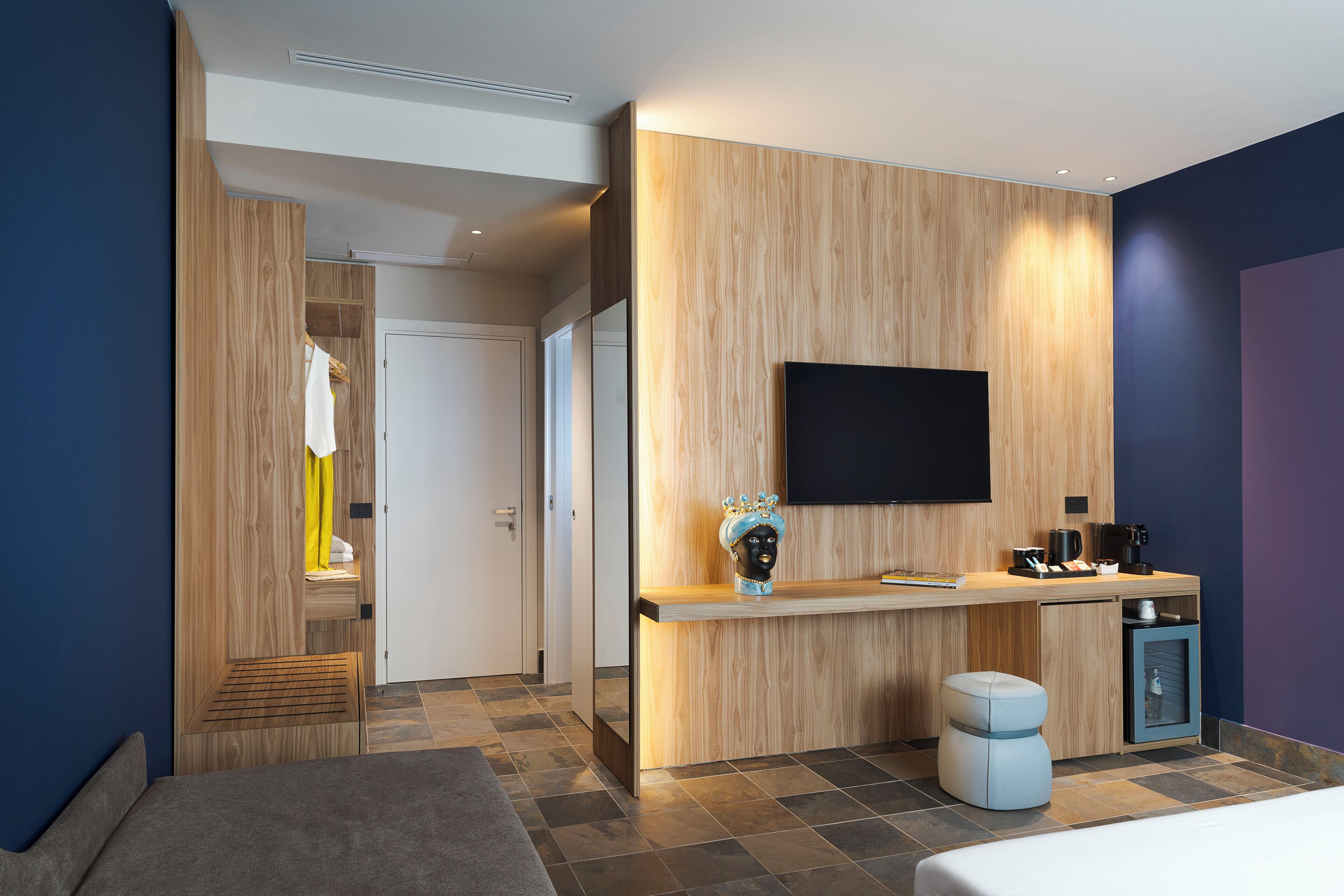 A modern hotel room with a wooden accent wall, a flat-screen TV, a bedside table with a coffee maker, and a small seating area.