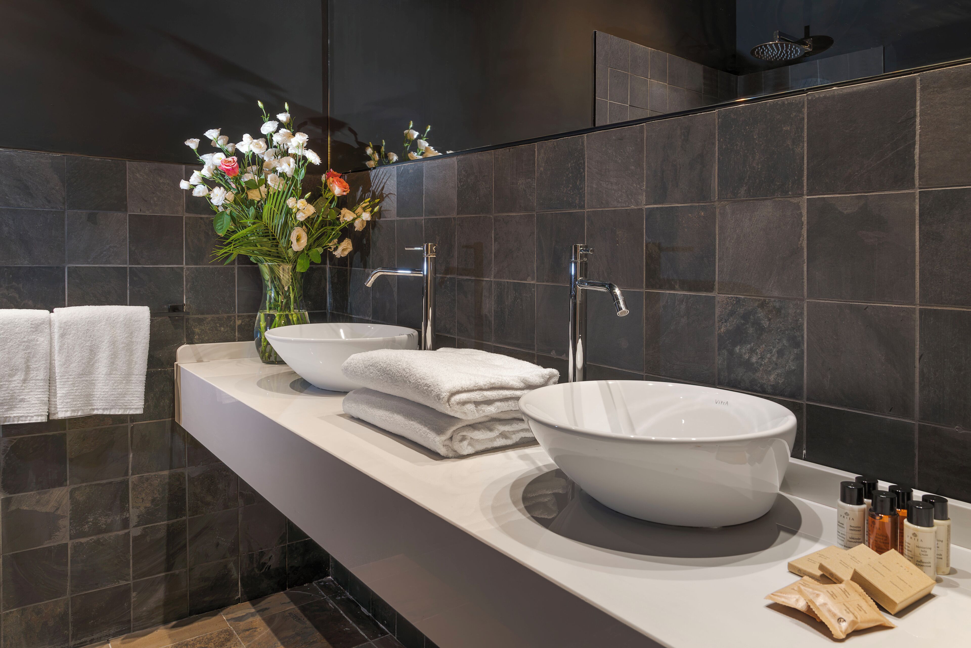 A modern bathroom with a large white sink, two towel stacks, a vase with flowers, and toiletries on the counter.