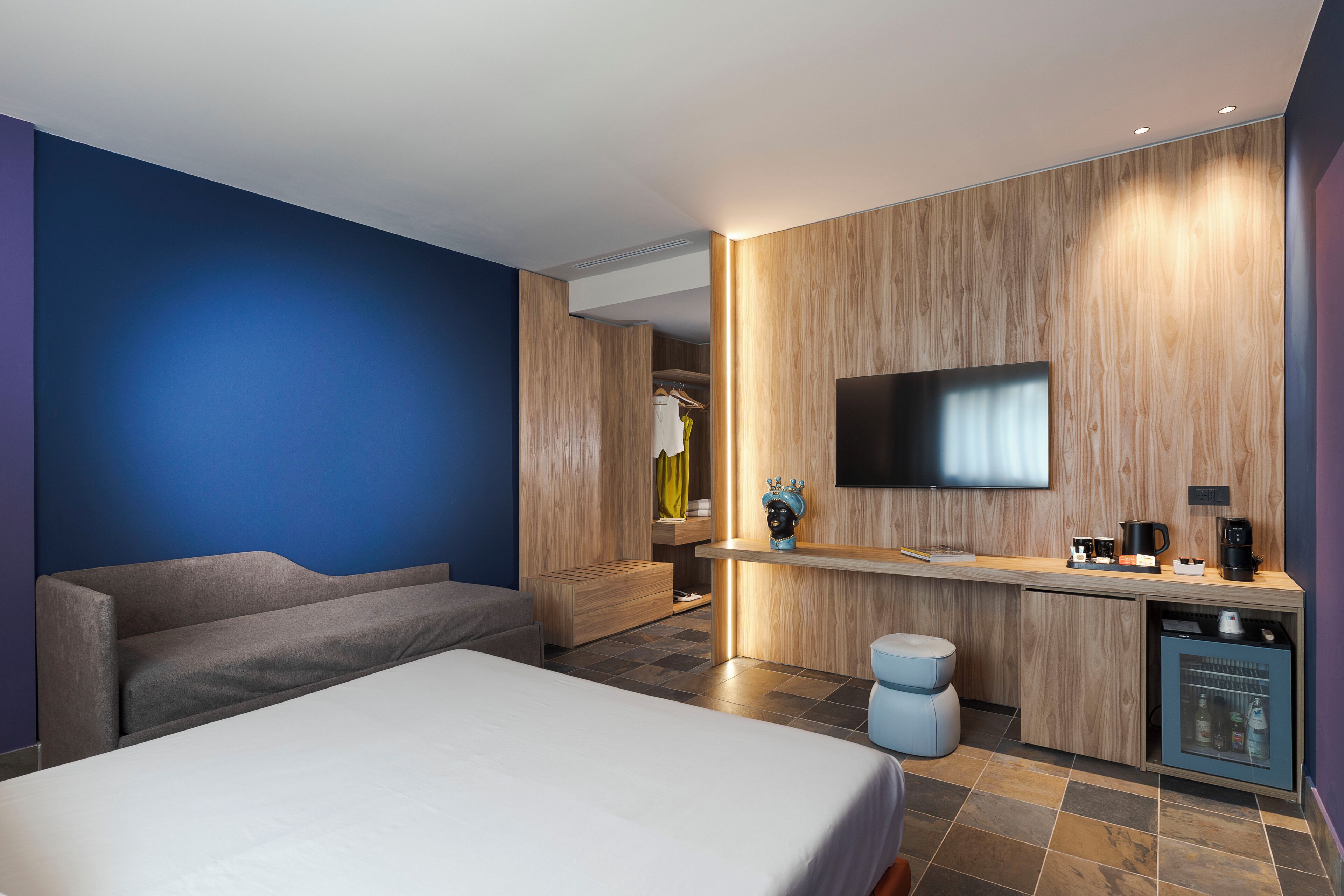 Ein modernes Hotelzimmer mit Bett, Sofa, Schreibtisch mit Flachbildfernseher und einer kleinen Küchenzeile.
