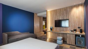 Ein modernes Hotelzimmer mit Bett, Sofa, Schreibtisch mit Flachbildfernseher und einer kleinen Küchenzeile.