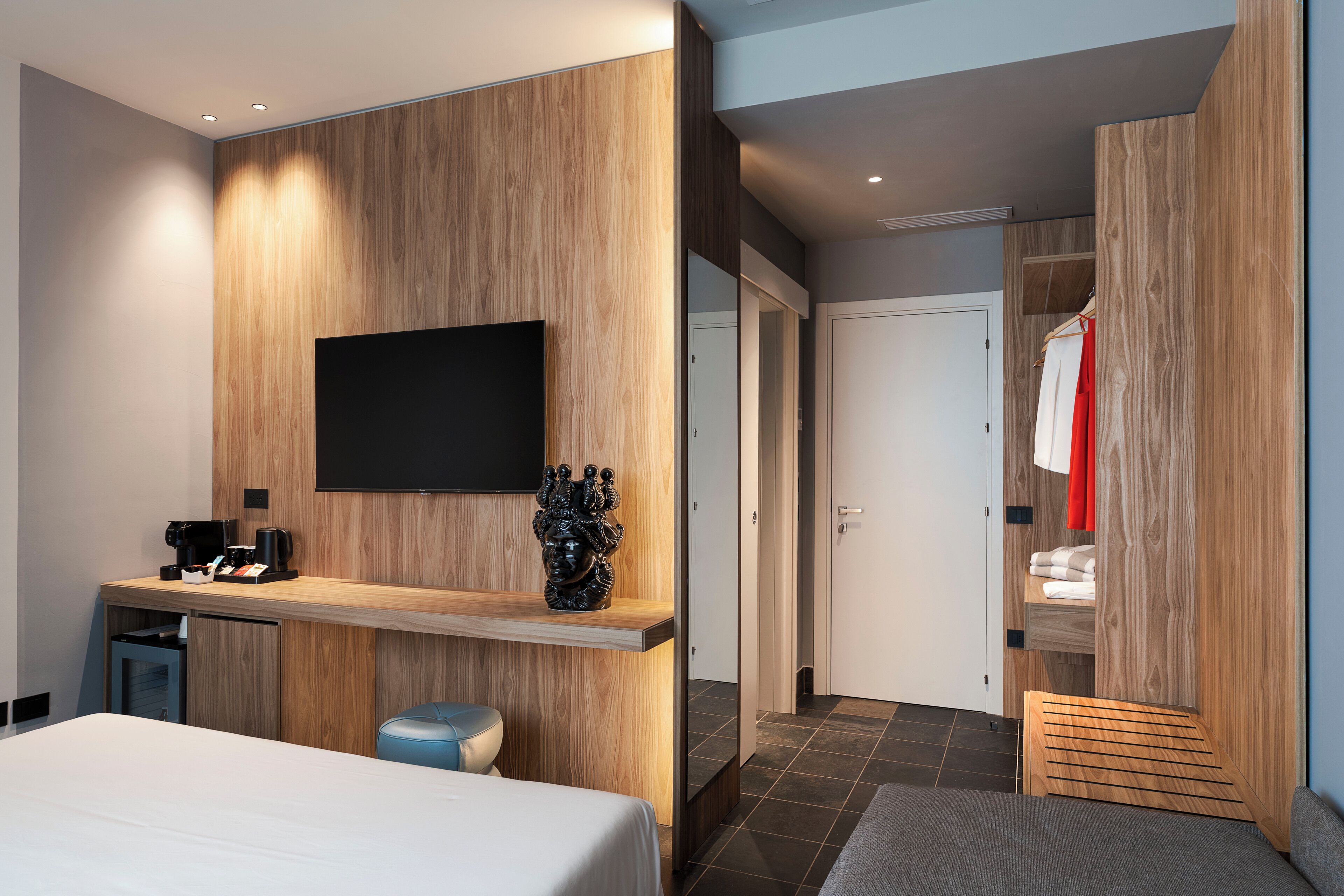 Ein modernes Hotelzimmer mit Bett, Schreibtisch mit Kaffeemaschine, Flachbildfernseher und Kleiderschrank mit Kleidung.
