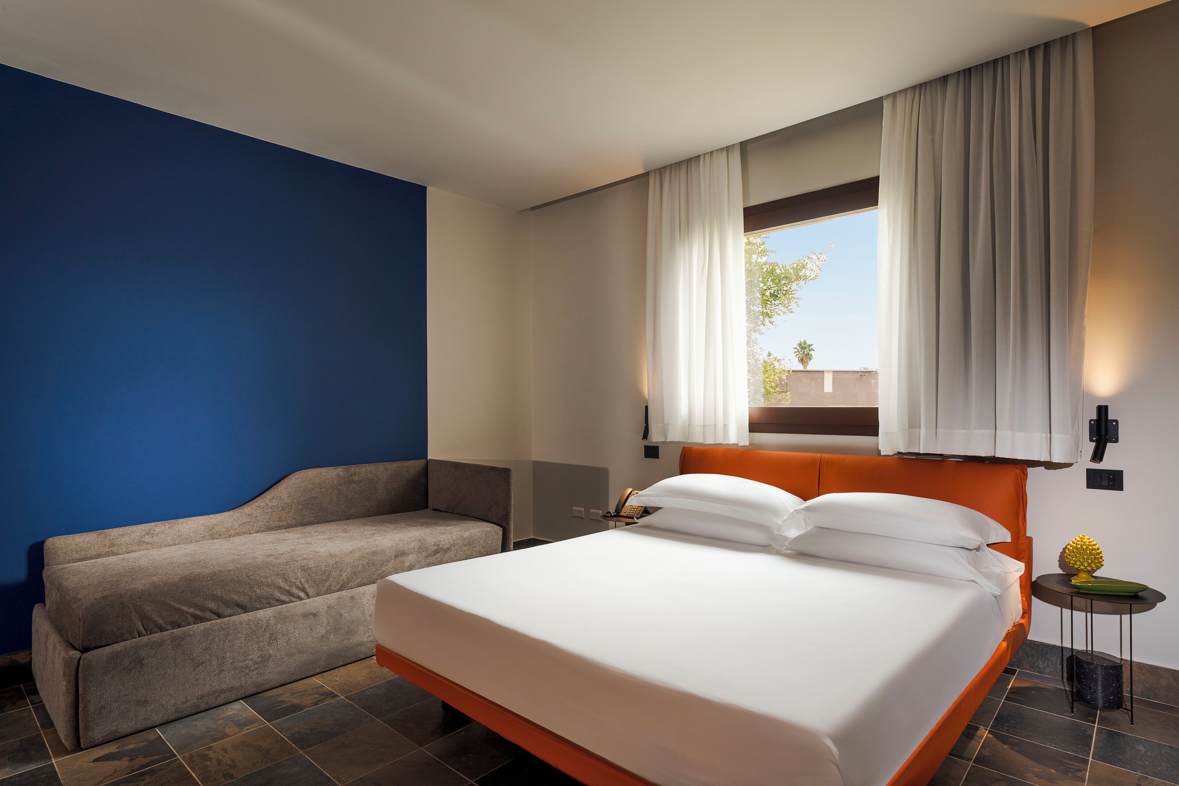 A hotel room with a bed, a sofa, a small table with a lamp, and a window with curtains.