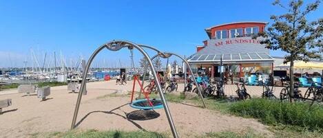 Ubicación frente a la playa con un columpio, un edificio identificado como “AM RUNDSTEIG” y bicicletas estacionadas cerca.