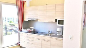 Cozinha compacta com armários de madeira clara, micro-ondas, máquina de café e janela com vista para um estacionamento.