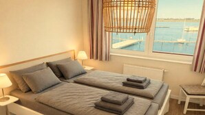 Cama de casal com roupa de cama e almofadas cinzas, mesa de cabeceira com abajur, e vista para uma marina pela janela.