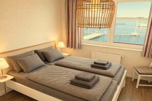 Ein Doppelbett mit grauer Bettwäsche und Kissen, ein Nachttisch mit Lampe und Blick auf einen Yachthafen durch ein Fenster.