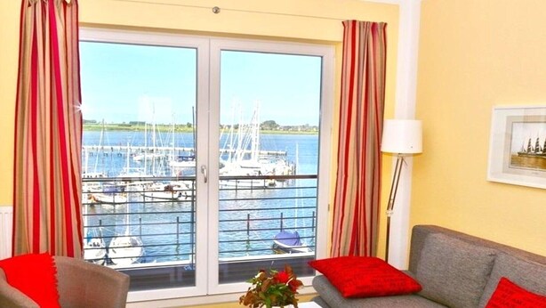 Quarto com sofá, poltrona, mesa e vista para uma marina com barcos.