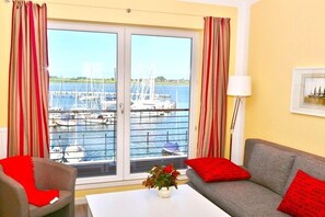 A room with a sofa, a chair, a table, and a view of a marina with boats. - Apartment am Südstrand in Fehmarn-Burgtiefe (Fehmarn OT Burgtiefe-Südstrand)