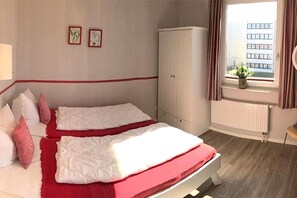 A hotel room with two beds, a wardrobe, a window with curtains, and a view of buildings outside. - Ferienwohnung in Fehmarn-Burgtiefe (Fehmarn OT Burgtiefe-Südstrand)