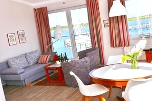 A room with a sofa, a round table, and chairs, a vase with flowers, and a view of the sea through large windows. - Ferienwohnung in Fehmarn-Burgtiefe (Fehmarn OT Burgtiefe-Südstrand)