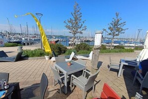 Eine Terrasse im Freien mit Tischen und Stühlen, eine gelbe Flagge und ein Yachthafen mit Booten im Hintergrund.