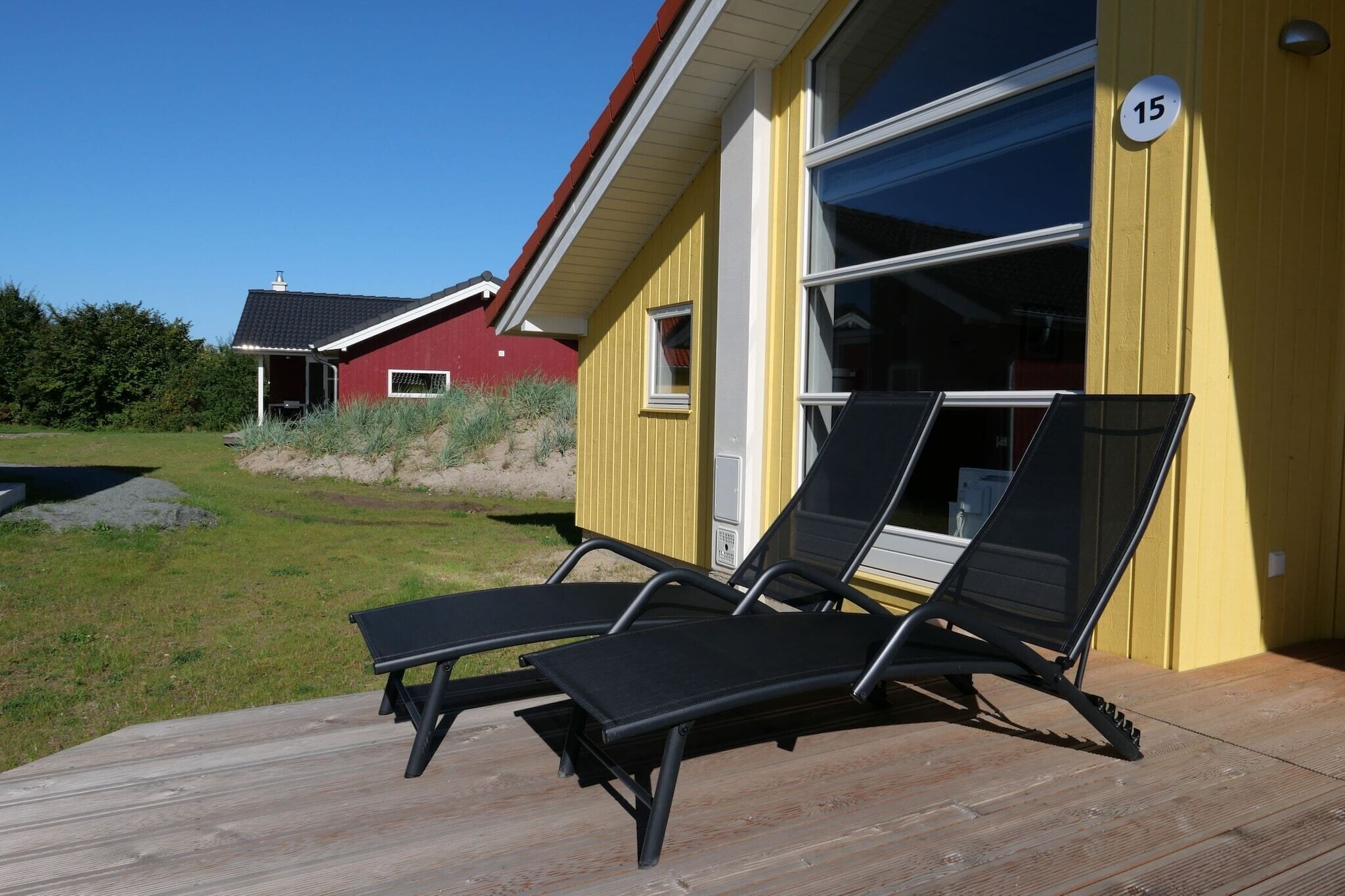 Un bâtiment jaune portant le numéro 15, une terrasse avec deux chaises longues noires et, au fond, un bâtiment rouge.