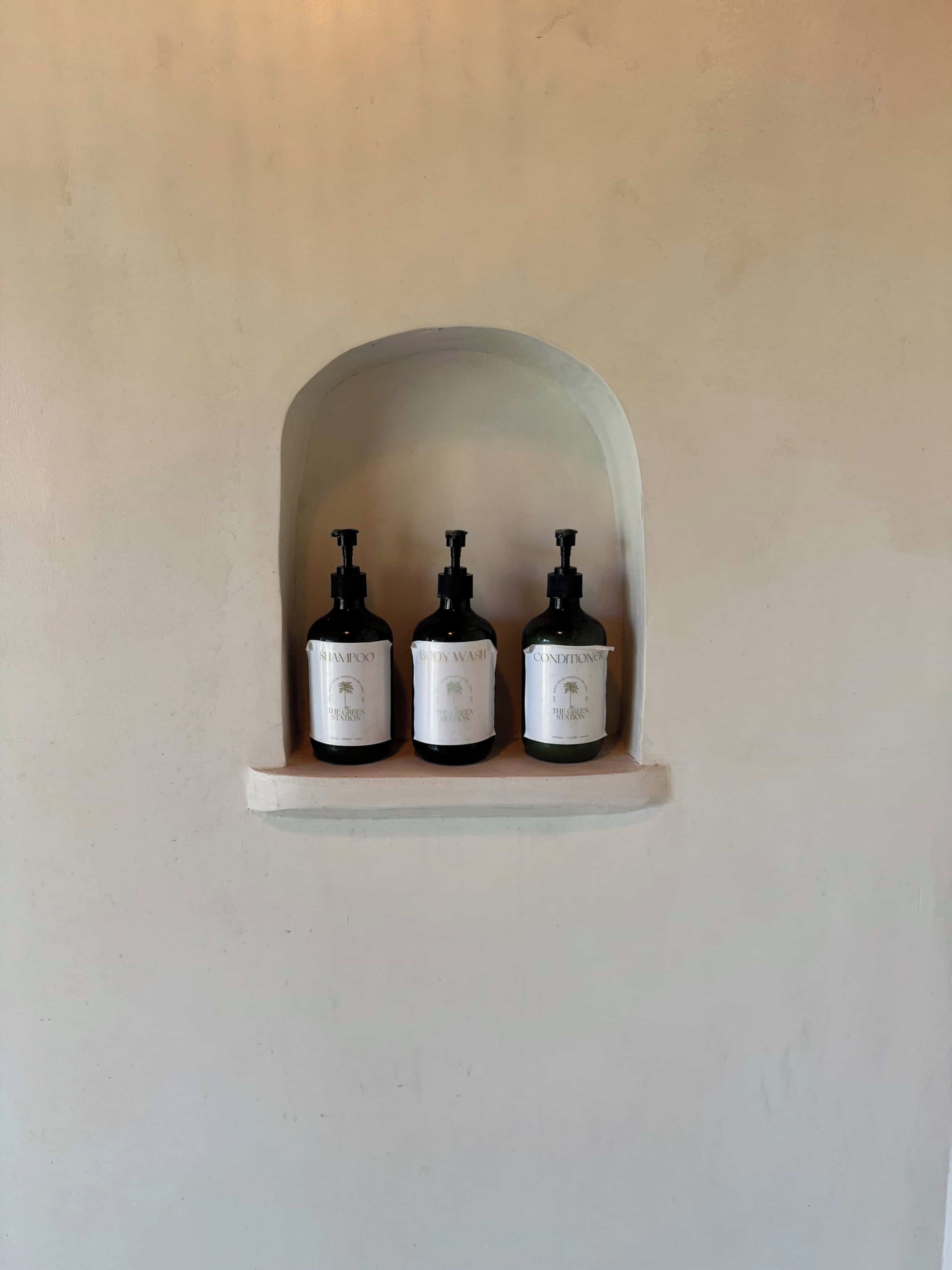 Une étagère avec trois bouteilles de produits de la marque The Green Station : un shampooing, un gel douche et un après-shampooing.