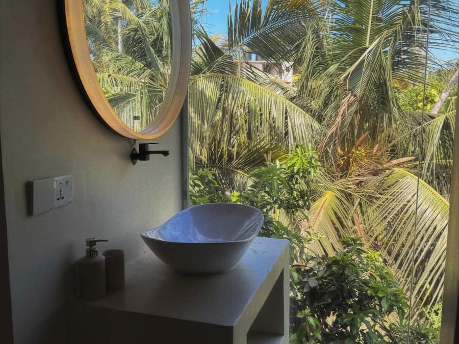 A bathroom with a large oval mirror, a white sink, and a view of tropical foliage through a window.