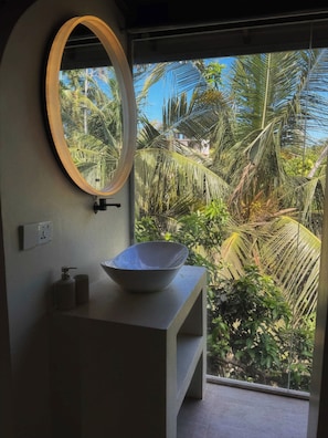 A bathroom with a large oval mirror, a white sink, and a view of tropical foliage through a window.