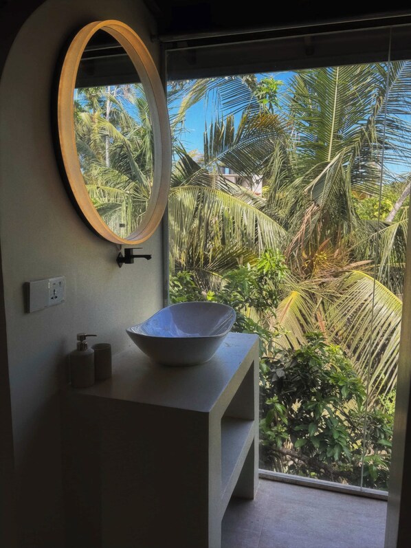 Un baño con un espejo ovalado grande, un lavabo blanco y vistas a follaje tropical a través de una ventana.
