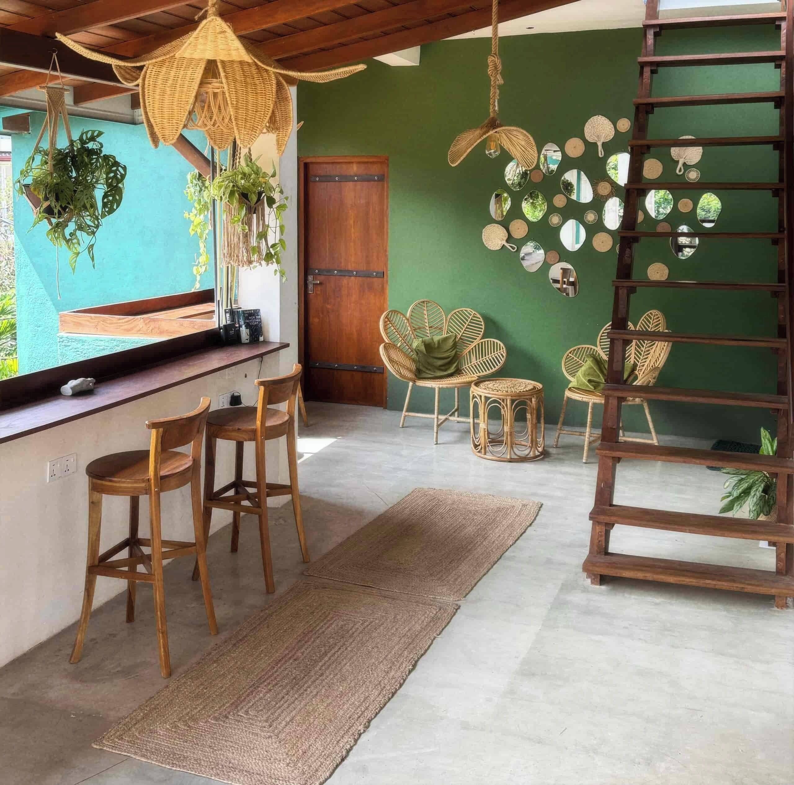 Una habitación con techo de madera, pared verde, puerta de madera y una lámpara colgante. Hay dos sillas de madera, un tapiz tejido y una pequeña mesa redonda con una planta.