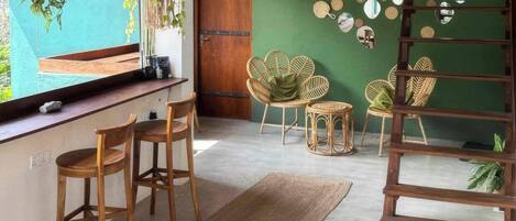 Una habitación con techo de madera, pared verde, puerta de madera y una lámpara colgante. Hay dos sillas de madera, un tapiz tejido y una pequeña mesa redonda con una planta.