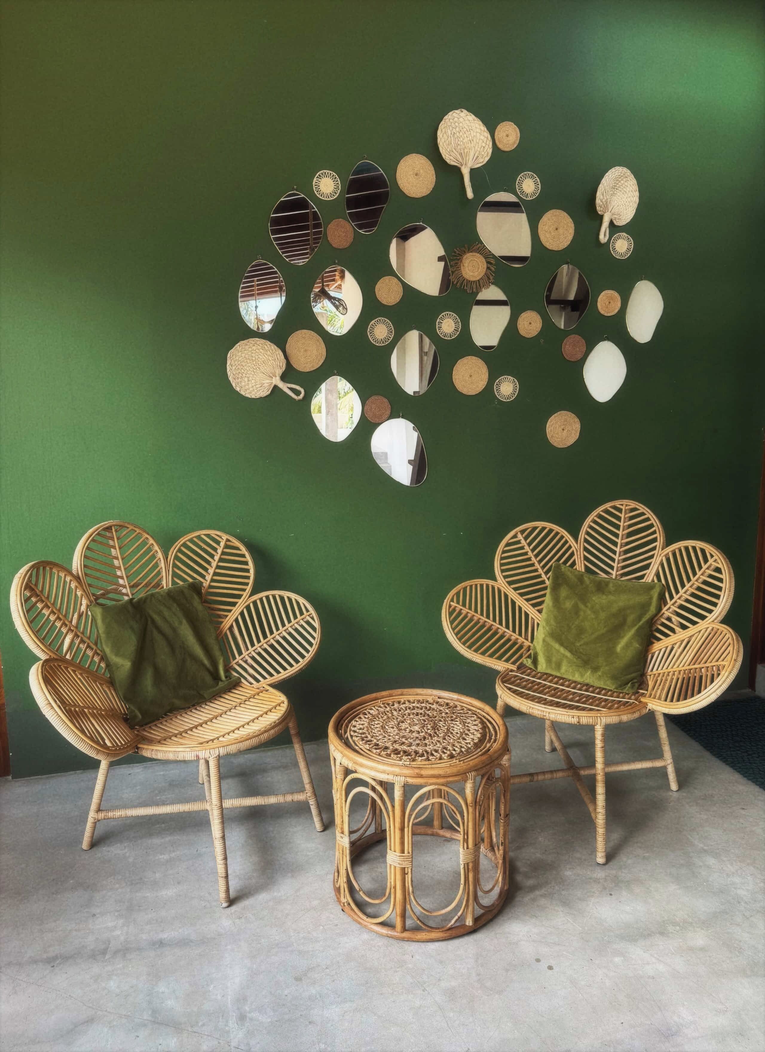 Twee rieten stoelen met groene kussens en een kleine ronde rieten tafel tegen een groene muur.