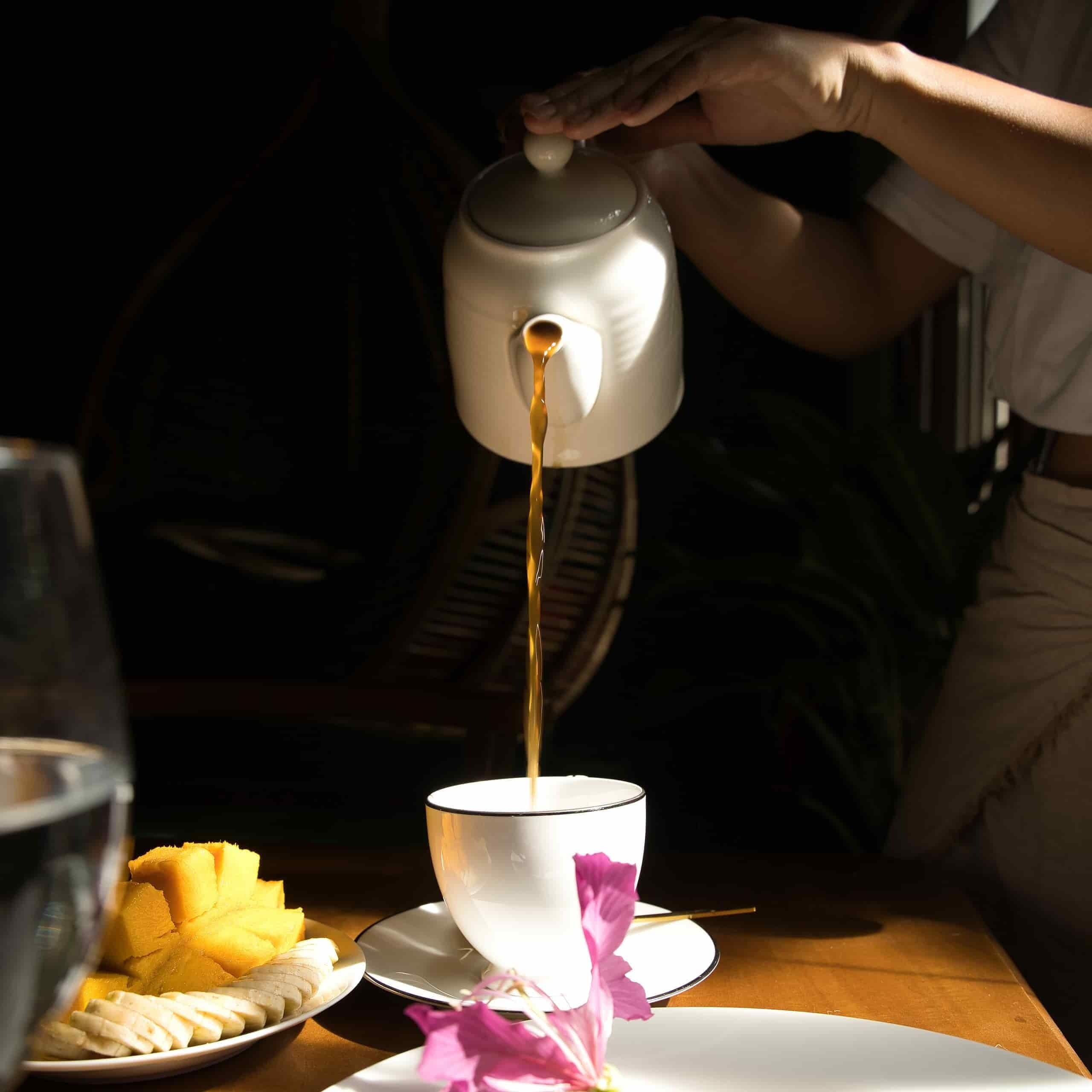 A person pouring tea into a cup on a table with a plate of food and a glass of water.