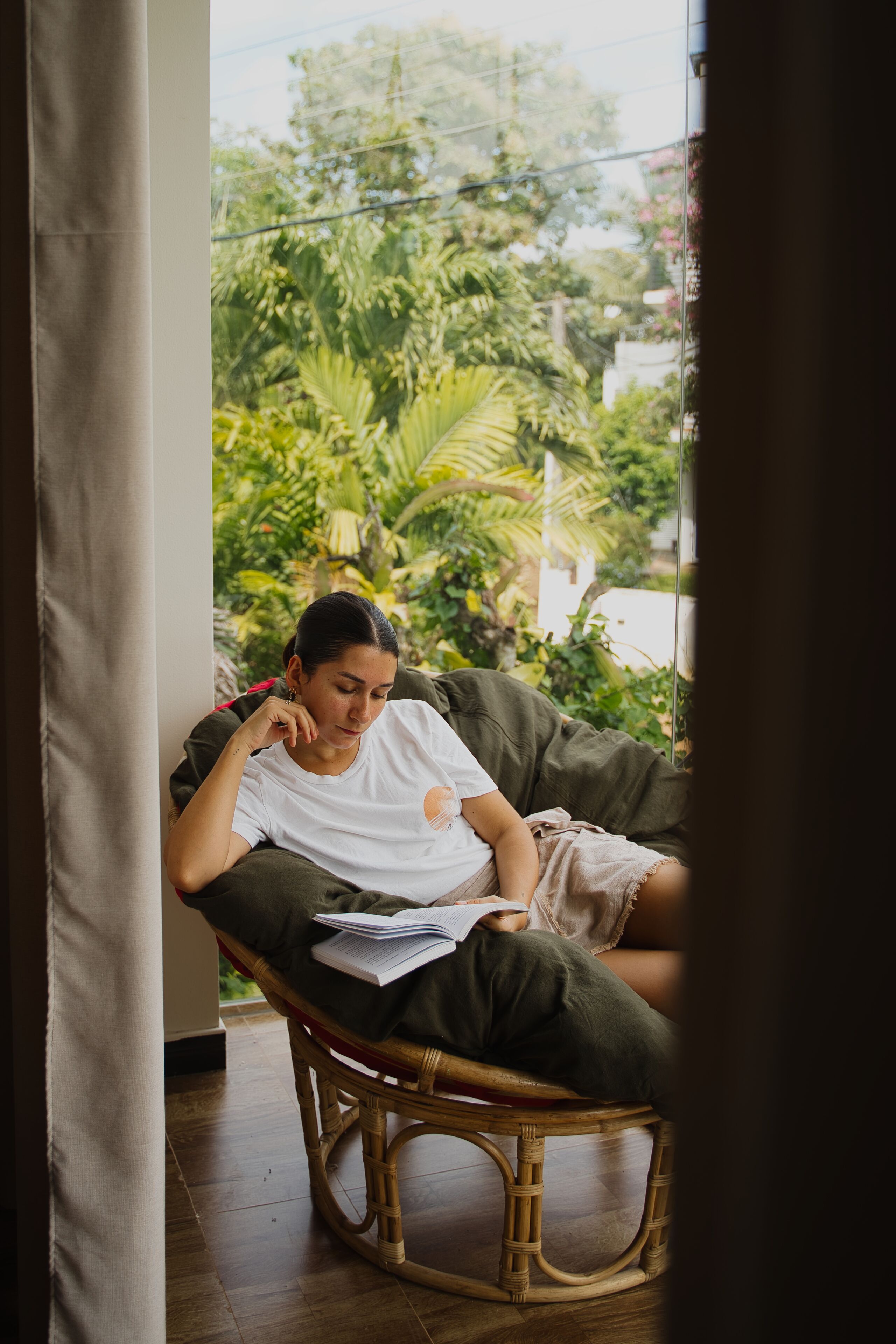 A person seated on a wicker chair with a book, in a room with a view of greenery outside.
