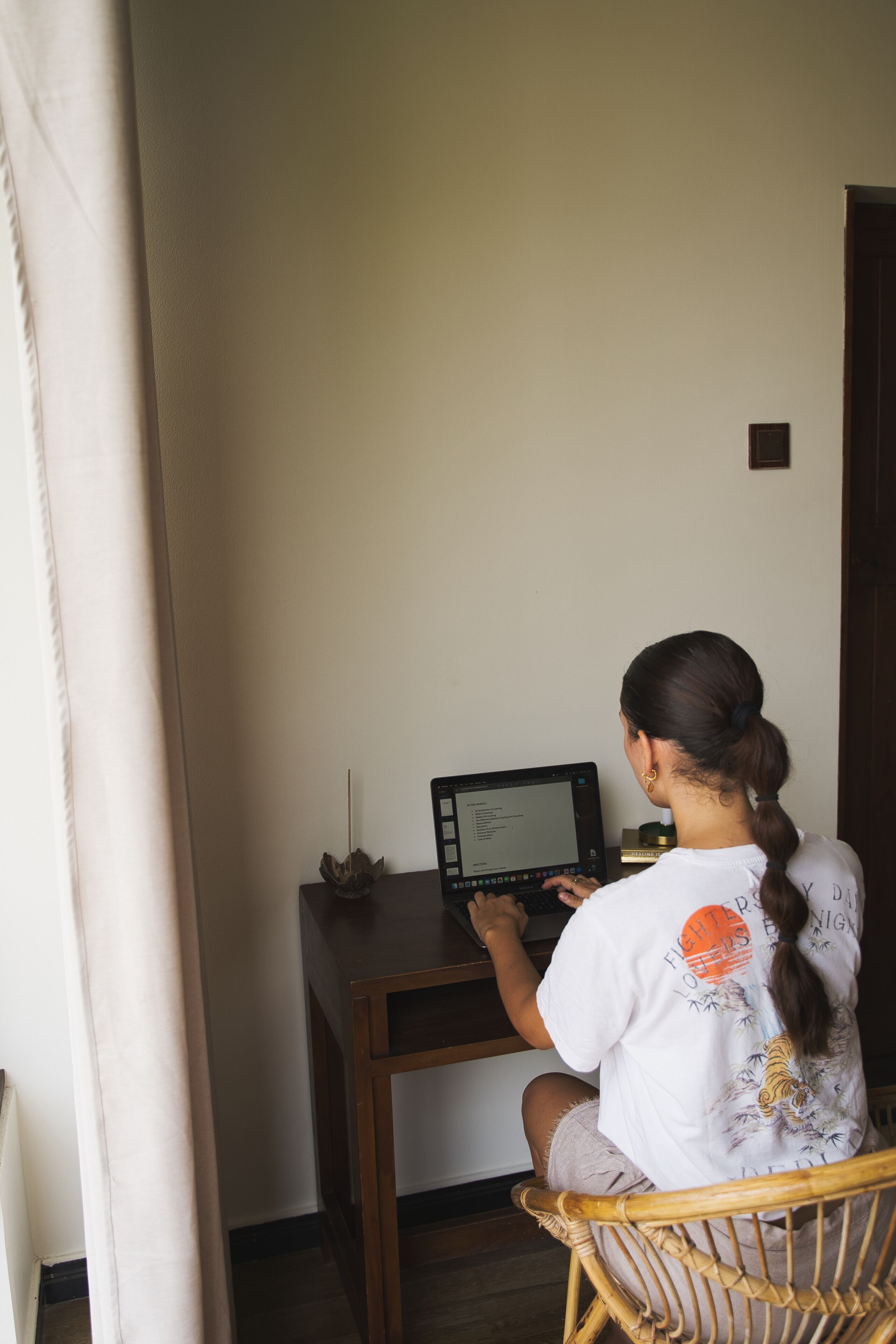 Una persona sentada en un escritorio de madera con una computadora portátil, en una habitación con una pared lisa y una puerta al fondo.