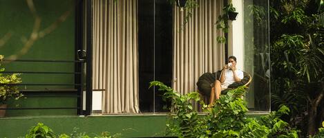 A person sitting on a chair by a window with vertical blinds, surrounded by greenery.