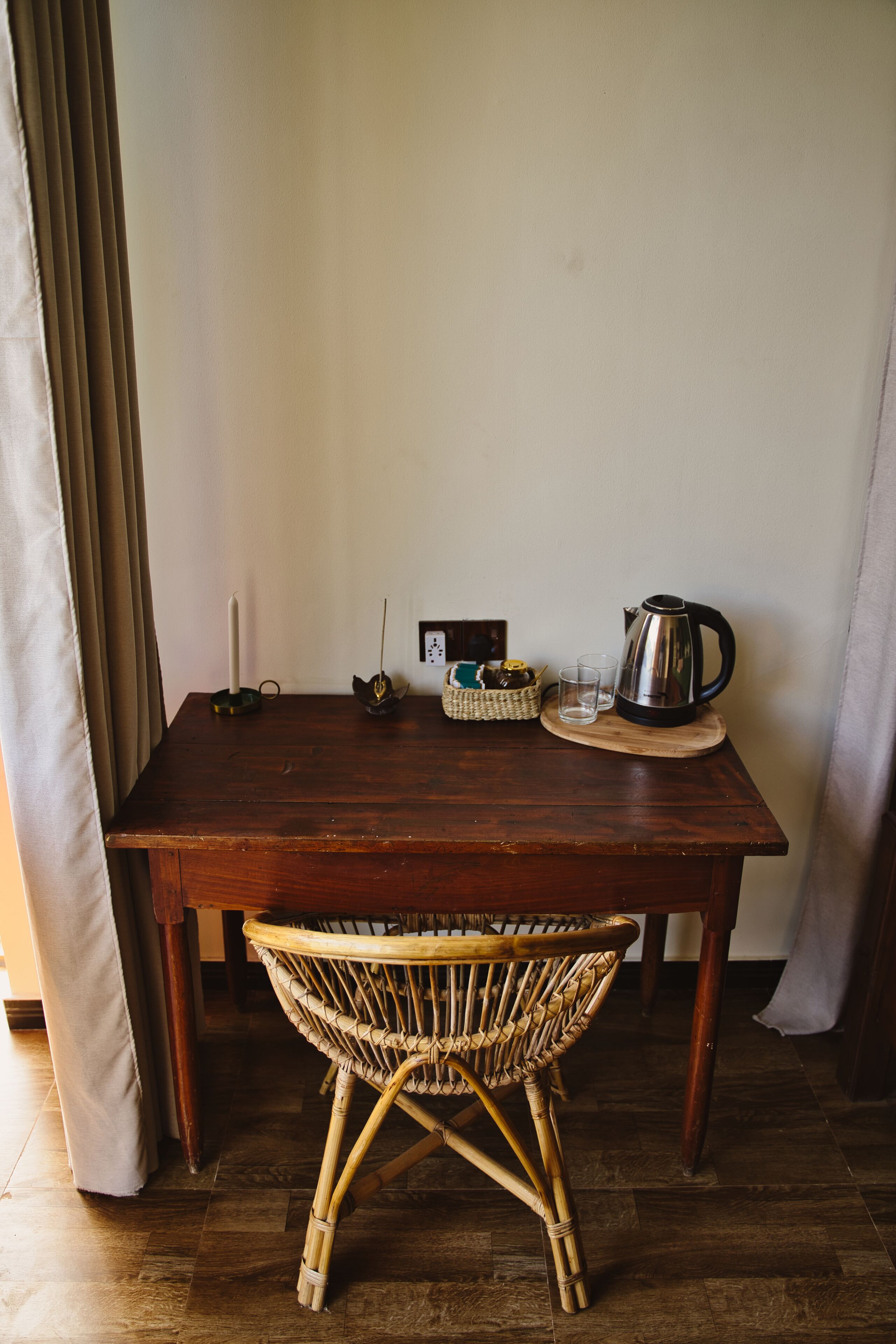 A wooden table with a wicker chair in front, a candle, a kettle, and a basket on the table.