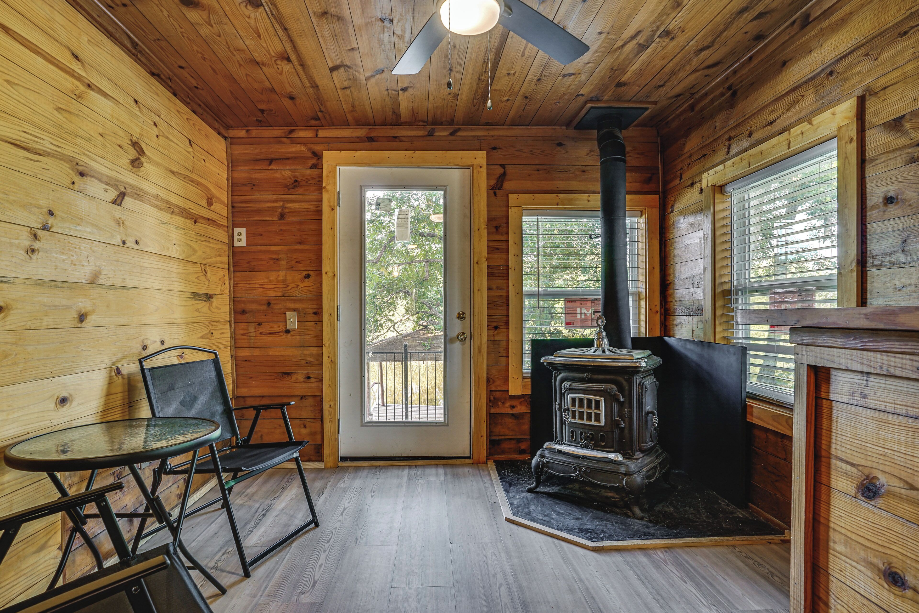 A wooden cabin interior with a door, a table with chairs, and a wood-burning stove.