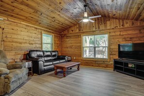 A log cabin interior with a leather sofa, a wooden coffee table, a television on a stand, and a ceiling fan. - Boat Dock & Ramp! Brazos River Couple’s Retreat (Granbury)