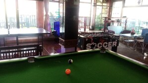 Una mesa de billar con superficie de fieltro verde y una bola blanca, una mesa de fútbol de mesa identificada como 'SOCCER', y un área de estar con mesas y sillas.