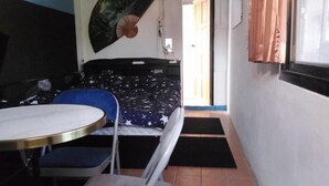 Una habitación compacta con cama, una mesita, una silla y un ventilador en la pared.