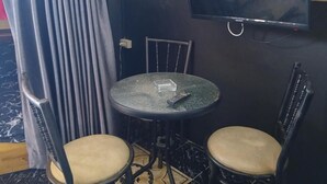 Um quarto pequeno com televisão na parede, duas cadeiras, uma mesa e uma cortina.