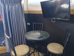 Pieni huone, jossa on seinälle asennettu televisio, kaksi tuolia, pöytä ja verho.
