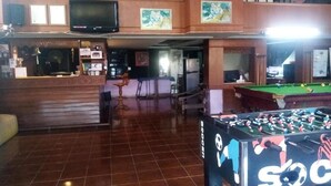 Um espaço amplo e interno, com mesa de pebolim, mesa de bilhar e uma televisão sobre uma plataforma de madeira.