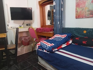 Quarto com cama, escrivaninha, cadeira, televisão e espelho.