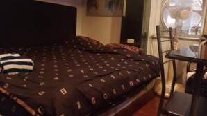 Ein Bett mit einem gemusterten Überwurf, ein Stuhl, ein Tisch und ein Wandventilator.