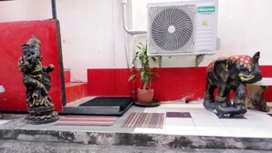 Un mur rouge et blanc orné d’une petite statue d’une divinité, d’une plante en pot et d’un ventilateur fixé au mur.