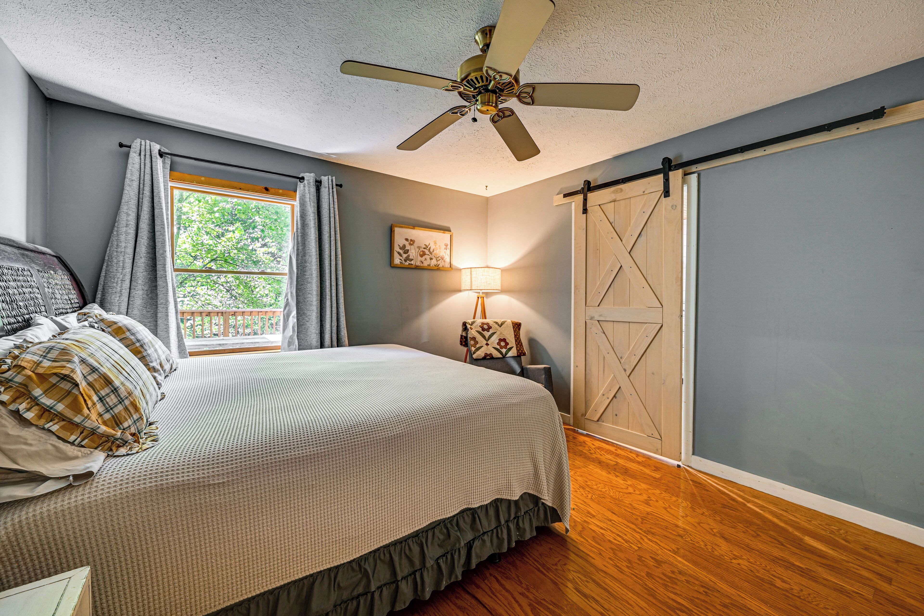 A bedroom with a bed, a ceiling fan, a window with curtains, a wall-mounted lamp, and a sliding barn door.