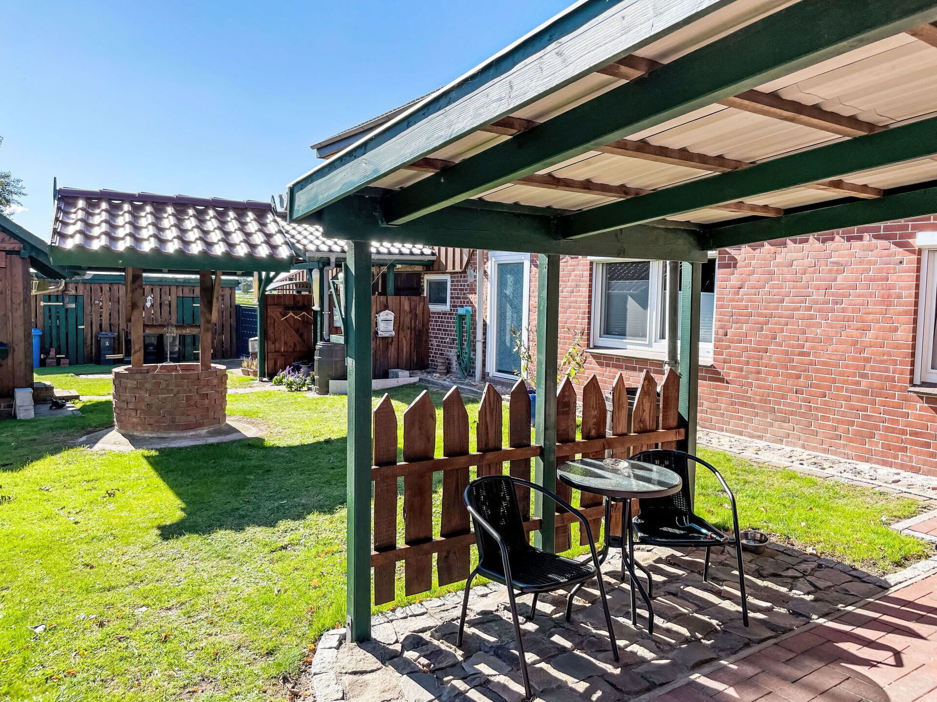 A covered patio area with two metal chairs and a small table, a brick barbecue pit, and a wooden fence.