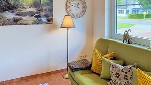 Ein Wohnzimmer mit einem grünen Sofa, einer Wanduhr, einer Stehlampe und einem Gemälde von einem Waldlauf.