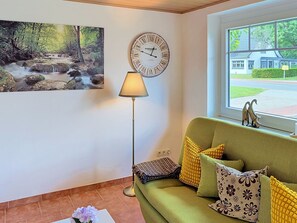 Ein Wohnzimmer mit einem grünen Sofa, einer Wanduhr, einer Stehlampe und einem Gemälde von einem Waldlauf.