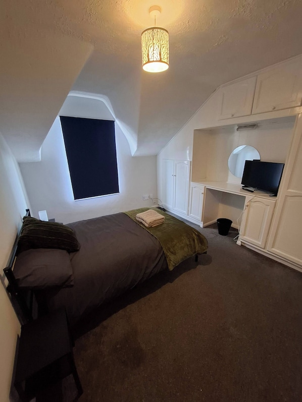 A bedroom with a bed, a desk with a computer, a television, and a window.