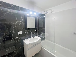 Un baño moderno con pared de mármol, un lavamanos blanco, un espejo grande y una ducha con mampara de vidrio.