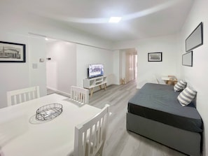 Una sala de estar moderna y bien iluminada, con un sofá gris, una mesa de comedor blanca y un televisor sobre un mueble blanco.